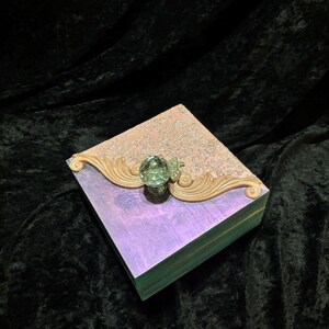 May include: A small, square wooden box with a pink and purple iridescent finish. The box has a decorative gold trim and a clear crystal knob.