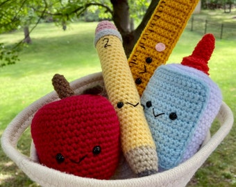 May include: A white crocheted basket filled with crocheted school supplies. The supplies include a red apple, a yellow pencil, a blue glue bottle, and a yellow ruler. Each item has a cute face.