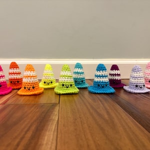 May include: A collection of eleven small, crocheted traffic cones in various colors including pink, orange, yellow, lime green, teal, blue, purple, and lavender. Each cone features white horizontal stripes and a stitched face with black eyes and a smile.