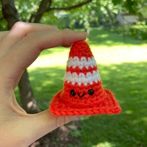 May include: A small, handmade crochet traffic cone featuring a bright orange base and body with a single horizontal white stripe. The cone has two small black plastic safety eyes and a stitched black V-shaped mouth, giving it a friendly facial expression.