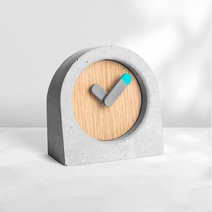 May include: A gray concrete clock with a wooden face and a blue and gray checkmark hand. The clock is a decorative piece for a home or office.