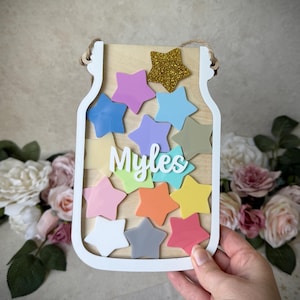 May include: A wooden jar-shaped sign with a white border, filled with colourful star cutouts. The name "Myles" is written in white script. A gold glitter star is at the top. The sign is held by a hand, with a floral background.