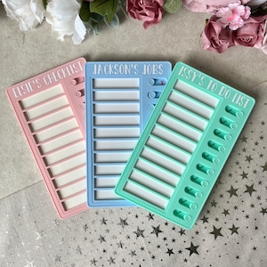 May include: Three colourful dry-erase chore charts with personalised names: Elsie's Checklist, Jackson's Jobs, and Issy's To Do List. Each chart has a white background with a light pink, light blue, or light green border. The charts are on a light grey surface with silver stars.