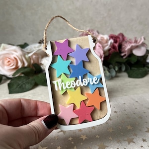 May include: A jar-shaped wooden ornament with a brown interior and white border, featuring colourful star cutouts. The name "Theodore" is written in white script. A twine loop is attached for hanging.