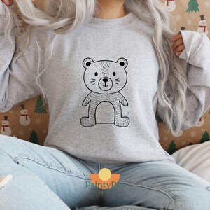 May include: A light gray sweatshirt featuring a black and white cartoon bear design. The bear has a simple, outlined style with a round head, small ears, and a smiling face. The sweatshirt is a long-sleeved, crew neck style.