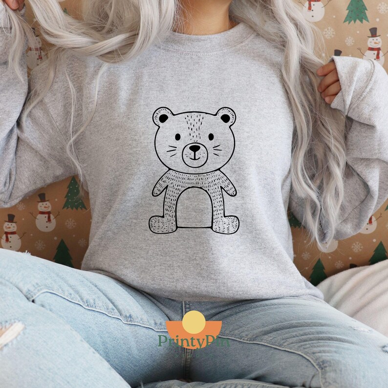 May include: A light gray sweatshirt featuring a black and white cartoon bear design. The bear has a simple, outlined style with a round head, small ears, and a smiling face. The sweatshirt is a long-sleeved, crew neck style.