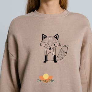 May include: A beige sweatshirt featuring a black and white fox graphic. The fox is stylized with a detailed body and tail. The PrintyPin Studio logo is at the bottom.