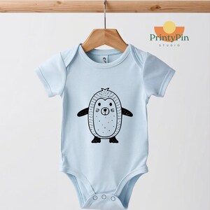 May include: A light blue baby bodysuit with a black and white penguin graphic. The penguin has a round body, small wings, and a smiling face. The bodysuit is hanging on a wooden hanger.
