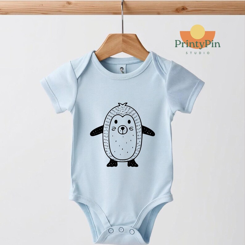 May include: A light blue baby bodysuit with a black and white penguin graphic. The penguin has a round body, small wings, and a smiling face. The bodysuit is hanging on a wooden hanger.