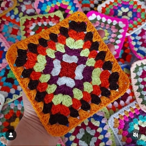 May include: A colourful crocheted granny square with a central motif of red, white and purple. The square is surrounded by a border of orange, black and green yarn.