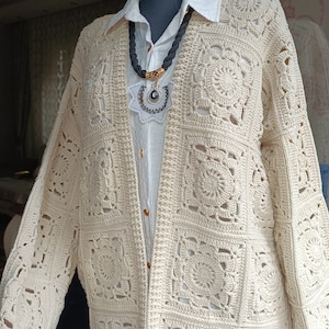 May include: A cream-colored crocheted cardigan with a square pattern. The cardigan is open in the front and has long sleeves. It is displayed on a mannequin with a white shirt underneath.
