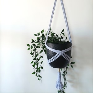 May include: A black hanging planter with a light blue macrame design and tassel, holding a green plant. The planter is suspended by a beige rope and light blue cords. The plant has long, cascading green leaves.