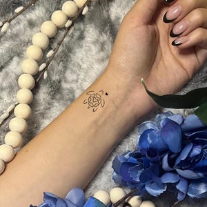 May include: A black line art turtle tattoo with a small heart on a wrist. The tattoo is on a light skin tone. The background includes a blue flower and a string of white wooden beads.