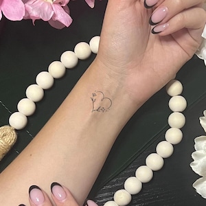 May include: A delicate, minimalist tattoo of a heart shape with floral accents on a wrist. The fine line art features a heart outline with leaves and small floral details. The skin tone is fair, and the tattoo is in black ink.