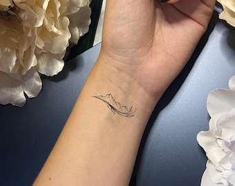 May include: A black line art tattoo of a mountain range with a leaf design on a wrist. The tattoo is small and minimalist, with fine lines and details. The skin tone is light.
