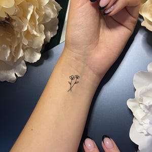 May include: A minimalist black outline tattoo of two small flowers on a wrist. The delicate design features two stems with heart-shaped petals, creating a simple yet elegant floral motif. The skin tone is fair, and the tattoo is freshly done.