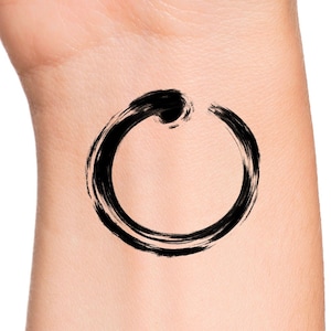 May include: A black temporary tattoo of an Enso circle, a symbol of Zen Buddhism, applied to the wrist. The brushstroke design features a thick, textured line forming a circle with an opening at the top. The tattoo is on a light skin tone.