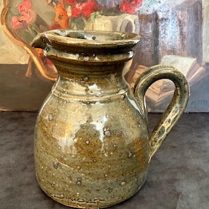 Könnte beinhalten: Handgefertigter, gesprenkelter Keramikkrug mit Ausguss und Henkel. Der Krug hat einen abgerundeten Körper und eine dunkelgrüne und braune Glasur. Der Griff ist gewölbt und der Ausguss leicht angewinkelt. Wahrscheinlich Vintage-Keramik.