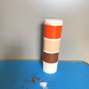 Könnte beinhalten: Ein weißer, orangefarbener, brauner und beiger Plastik-Gewürzbehälter mit Deckel. Der Behälter steht auf einer blauen Oberfläche mit Salz und Pfefferkörnern, die um ihn herum verstreut sind.