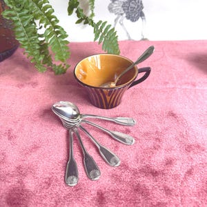 Peut inclure: Une tasse à thé vintage avec un intérieur marron et doré et une anse marron foncé, avec une cuillère en argent à l'intérieur. Une collection de cuillères en argent avec un motif de coquillage est disposée sur une surface en velours rose. Une fougère et un dessin décoratif sont en arrière-plan.