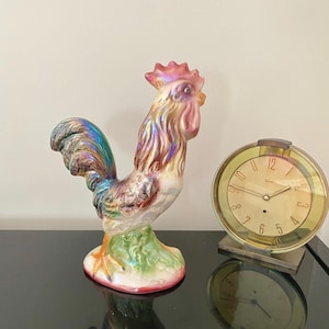 Puede incluir: Una figurita de gallo de cerámica con pintura iridiscente en tonos rosa, verde y marrón. El gallo está de pie sobre una base verde y mira hacia la derecha. Un reloj dorado con números romanos negros está en el fondo.