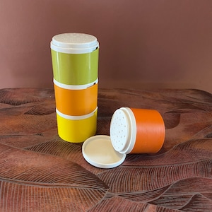 May include: A stack of three cylindrical plastic shakers in lime green, orange, and yellow, each featuring a white perforated lid. A separate orange shaker lies on its side next to a detached white circular lid, showing the perforated top design.