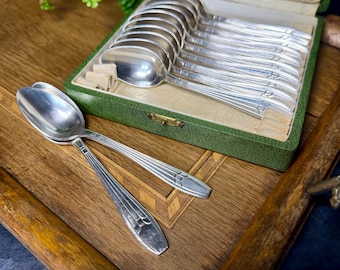 May include: A collection of silver spoons presented in a green velvet-lined box. Two spoons lie outside the box on a wooden tray. Each spoon features a rounded bowl and a decorative handle. The box is open, displaying the other spoons.