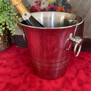 Könnte beinhalten: Silberner Champagnerkühler mit einer Flasche Champagner darin. Der Kühler hat zwei Griffe mit Ringen. Der Kühler steht auf einer roten Oberfläche.
