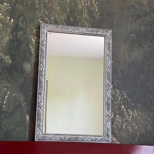 Può includere: Uno specchio da parete rettangolare con una cornice color argento caratterizzata da un intricato motivo in rilievo di fiori stilizzati e raggi solari su tutti e quattro i lati, che circonda una superficie in vetro chiara e riflettente.