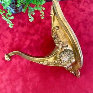Op de afbeelding: Een gouden metalen plankbeugel met sierlijke details en een gebogen arm. De beugel heeft een schelpvormig ontwerp en is in vintage stijl.