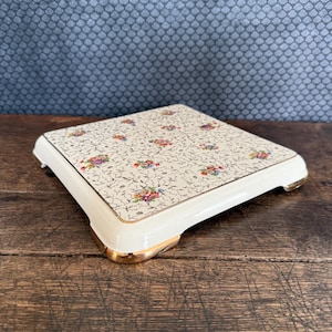 Peut inclure: Un plateau de service carré en céramique blanc cassé avec un motif floral et une bordure dorée. Le plateau présente un bord crème et des accents dorés sur les pieds. Le motif floral comprend des fleurs roses, violettes et jaunes.