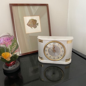 May include: A white ceramic mantel clock with gold trim and a round clock face. The clock is on a black surface next to a framed fish print and a decorative glass egg with flowers.