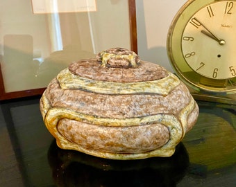 May include: A ceramic lidded jar with a brown and beige glaze. The jar has a decorative design of raised lines and a textured surface. The jar is sitting on a black surface.