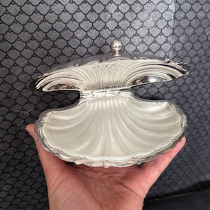 Op de afbeelding: Een zilverkleurige schelpvormige sieradendoos. Het scharnierende deksel is open en onthult een wit interieur. De doos heeft een gedetailleerd, geribbeld ontwerp en een kleine, ronde knop aan de bovenkant.