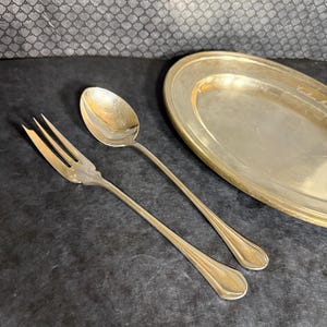 May include: Silverware set including a fork, spoon, and oval serving platter. The utensils have long, slender handles and a shiny finish. The platter has a slightly raised edge. The items are arranged on a dark surface.
