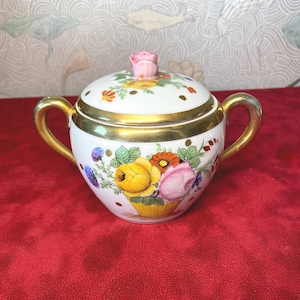 Può includere: Zuccheriera in porcellana bianca con coperchio e due manici. La ciotola è decorata con un motivo floreale in tonalità di rosa, giallo, arancio e blu. Il coperchio ha una rosa rosa in cima. La ciotola ha un bordo dorato.