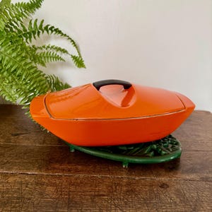 May include: A bright orange, oval-shaped enamelled cast iron casserole dish with a matching lid and a black handle. The dish is resting on a dark green, leaf-shaped metal trivet. The design features smooth, curved edges and a glossy finish.