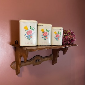 Puede incluir: Tres botes vintage blancos con diseños florales en un estante de madera. Los botes están etiquetados con "Farine", "Cafe" y "The".