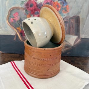 Puede incluir: Una mantequera de cerámica con tapa, con interior beige claro y exterior marrón rojizo. La tapa es de color beige claro. El plato está sobre un paño blanco con rayas rojas. El fondo incluye una pintura de flores.