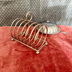 May include: A gold-colored metal toast rack with a circular handle and six curved slots. The rack sits on a rectangular base with small ball feet. A silver serving dish with a lid is in the background, resting on a red velvet surface.