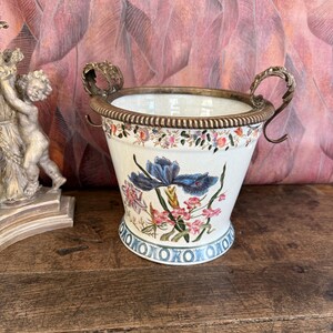 Peut inclure: Un pot de fleurs en céramique blanche avec un motif floral représentant des iris bleus et des fleurs roses. Le pot a des poignées en bronze et un bord décoratif. La base présente un motif géométrique bleu et blanc. Un objet décoratif pour la maison.