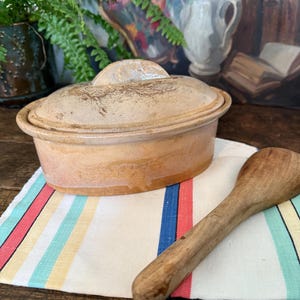 Może przedstawiać: Beżowe ceramiczne naczynie do zapiekania z pokrywką, spoczywające na pasiastym płótnie z czerwonymi, niebieskimi i żółtymi paskami. Drewniana łyżka leży obok naczynia. Tło obejmuje obraz i rośliny.