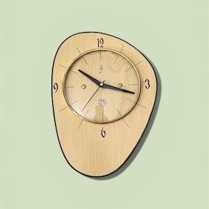 May include: A mid-century modern wall clock with a light wood grain face and a black outline. The clock has a gold-tone bezel and black hands. The numbers 3, 6, 9, and 12 are visible. The clock is mounted on a light green wall.