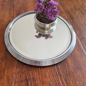 Könnte beinhalten: Ein versilberter Spiegel-Tablett mit einem kleinen silbernen Blumenfrosch, der einen Strauß lila und blauer Trockenblumen hält. Das Tablett reflektiert die Blumen und erzeugt eine dekorative Präsentation. Der Tablettdurchmesser beträgt ca. 25 cm.