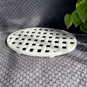 Puede incluir: Un salvamanteles ovalado de cerámica blanca con un diseño de celosía tejida. Este accesorio de cocina está diseñado para proteger las superficies de los platos calientes. El salvamanteles se muestra sobre una superficie gris estampada.
