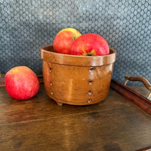 Può includere: Una ciotola in ceramica marrone con tre mele rosse all'interno e una mela accanto. La ciotola ha piccoli fori sui lati e poggia su un vassoio di legno. Le mele sono rosse con sfumature gialle.