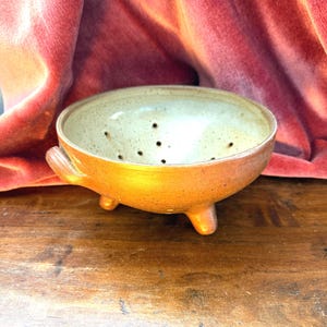 Puede incluir: Un cuenco de cerámica redondo para bayas con un interior crema moteado y un exterior de color marrón anaranjado cálido. El cuenco presenta pequeños orificios de drenaje en la base, dos pequeñas asas laterales y se apoya sobre tres patas cortas.