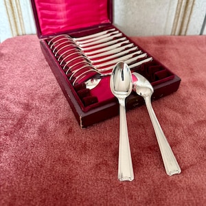 May include: A set of silver spoons displayed in a burgundy velvet-lined box. The spoons have a classic design with a rectangular handle and a decorative floral pattern at the base. The box is open, revealing the spoons arranged neatly.