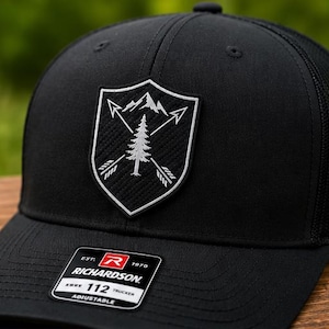 May include: Black trucker hat with a shield-shaped patch featuring a mountain range, arrows, and a tree. The patch is white and gray. The hat has a mesh back and a curved brim. The tag reads "RICHARDSON 112 TRUCKER ADJUSTABLE."