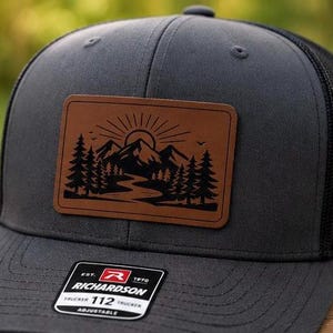 May include: Charcoal gray trucker hat with a black mesh back and a brown leather patch featuring a mountain and river scene. The hat has an adjustable strap and a Richardson 112 Trucker label.
